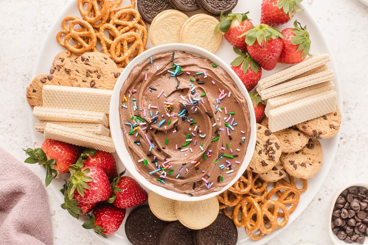 A white plate with strawberries, wafer cookies, pretzels, chocolate chip cookies, and sandwich cookies around a bowl of chocolate dip topped with colorful sprinkles.