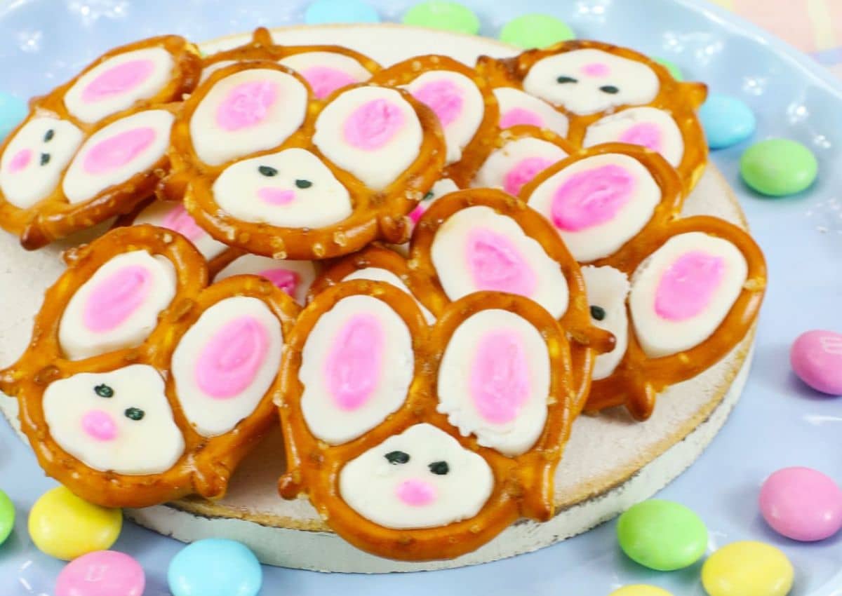 A plate of pretzel snacks decorated with white and pink candy to look like bunny faces, surrounded by colorful round candies.