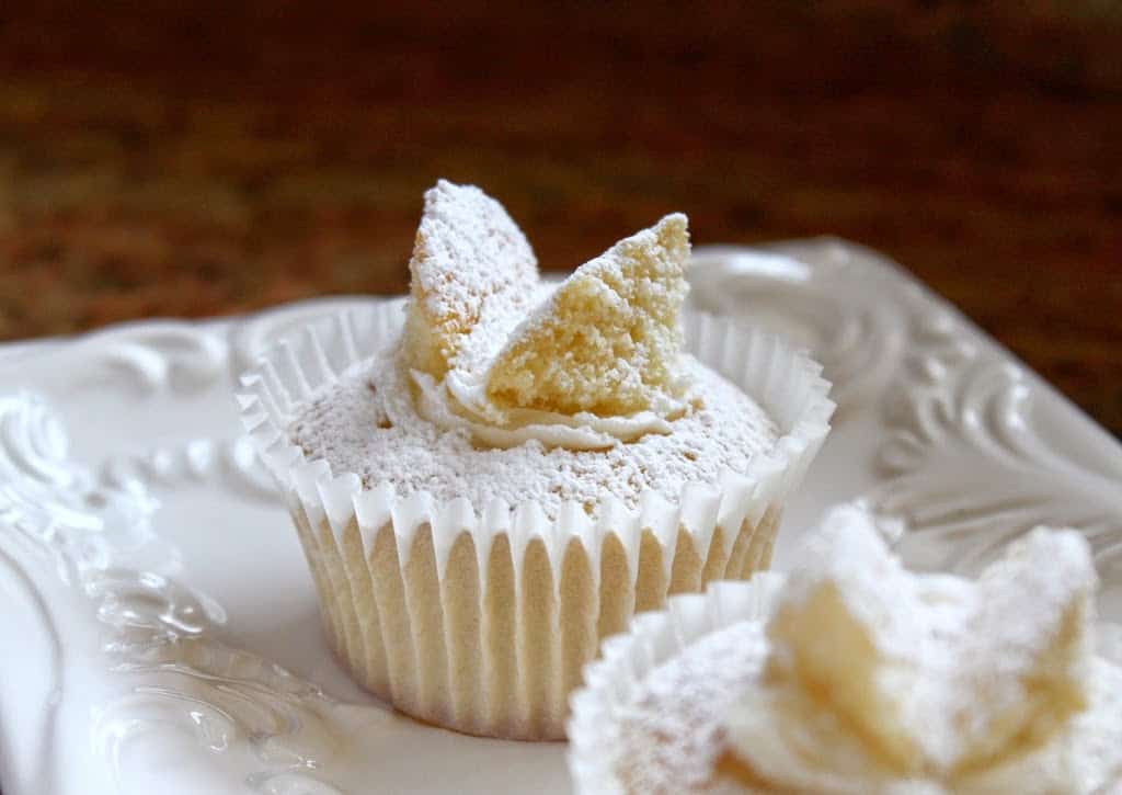 A vanilla cupcake with powdered sugar and two cake pieces on top, resembling butterfly wings, sits on a white decorative plate.