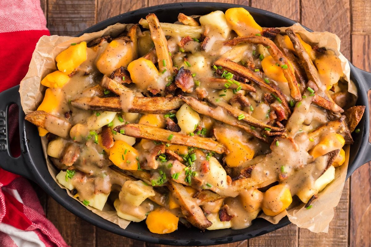 A dish of poutine with French fries, cheese curds, brown gravy, and chopped herbs in a black oval serving dish on a wooden table.