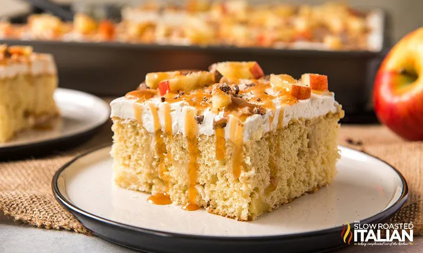 A slice of yellow cake topped with whipped cream, diced apples, caramel sauce, and nuts on a white plate, with more cake in the background.