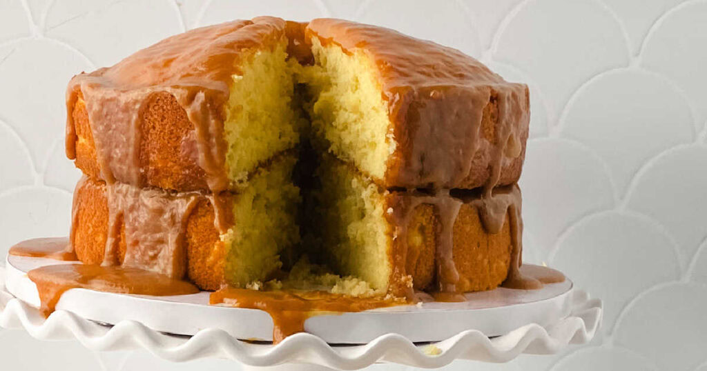 A two-layer yellow cake with caramel icing, sliced to show the inside, sits on a white cake stand against a white background.