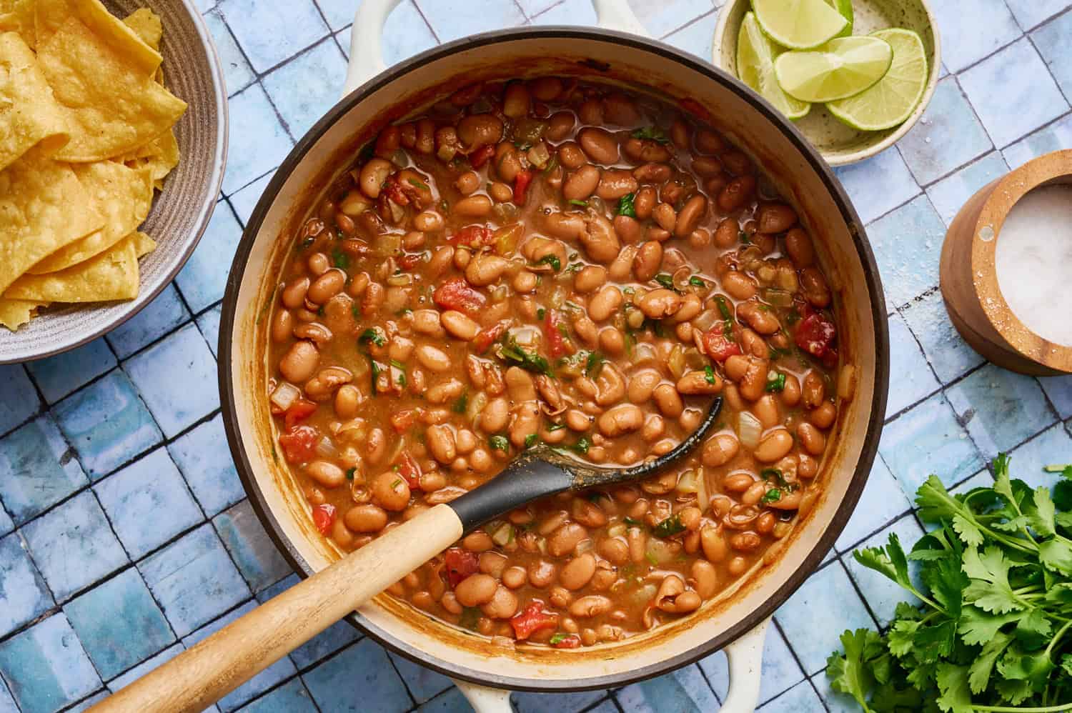 Pot of cooked pinto beans with a ladle, surrounded by tortilla chips, lime wedges, salt, and fresh cilantro on a tiled surface.