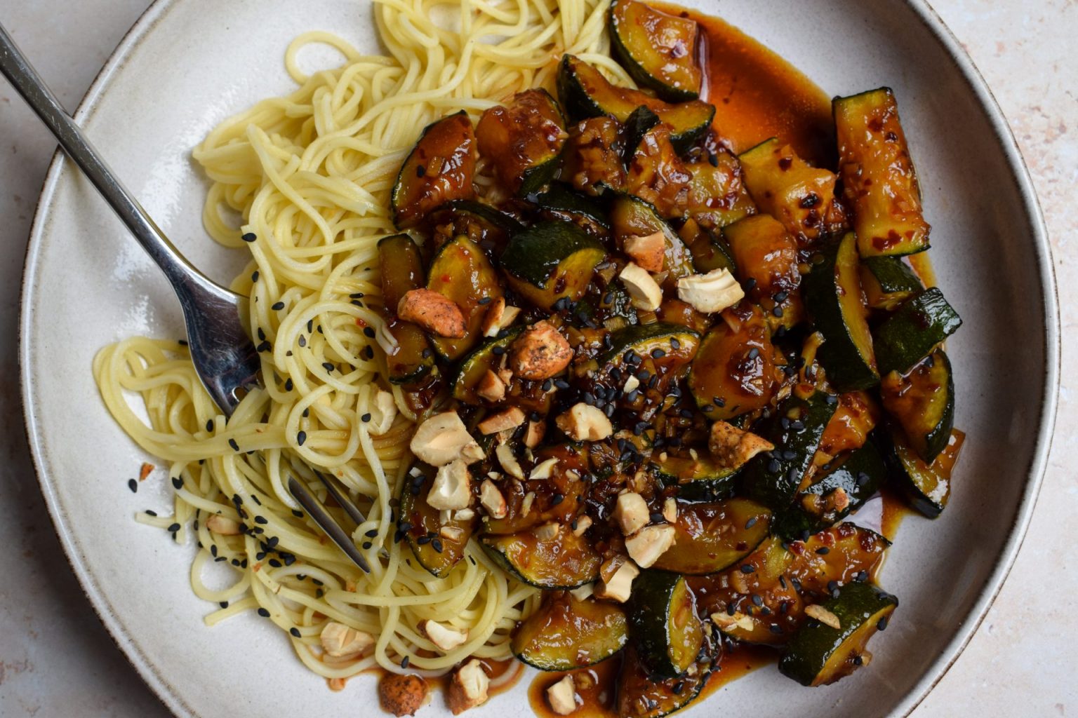 Plate of noodles topped with stir-fried zucchini in a dark sauce, garnished with chopped cashews and black sesame seeds, with a fork on the side.