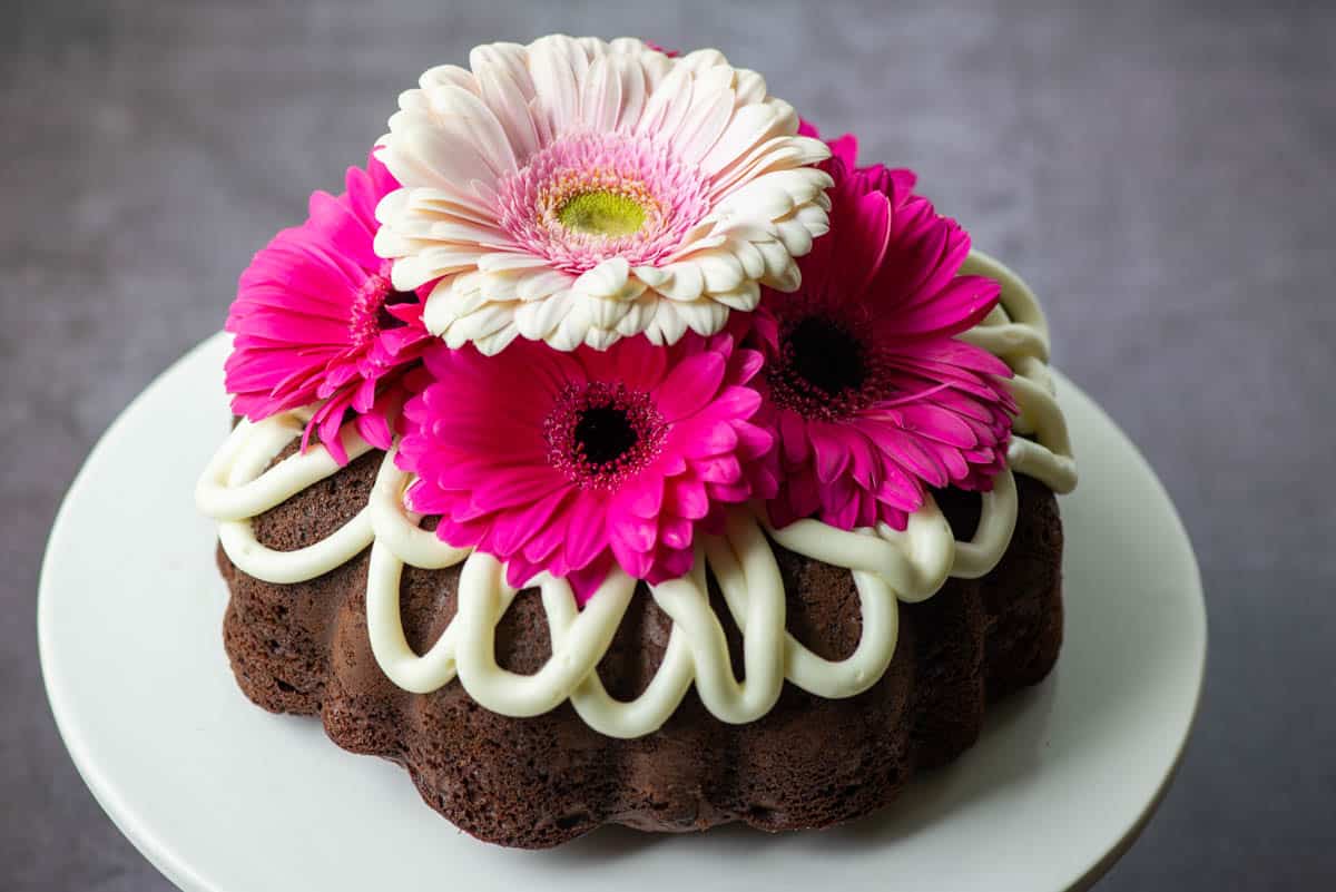 A chocolate bundt cake topped with white icing and decorated with pink and white gerbera daisies, displayed on a white cake stand.