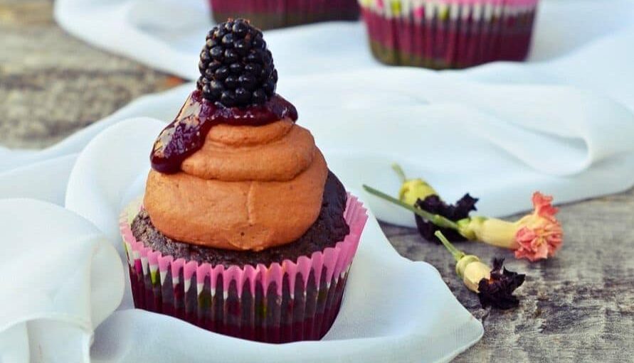 Chocolate cupcake with chocolate frosting, berry sauce, and a blackberry on top, in a pink wrapper, placed on a white cloth with small flowers nearby.