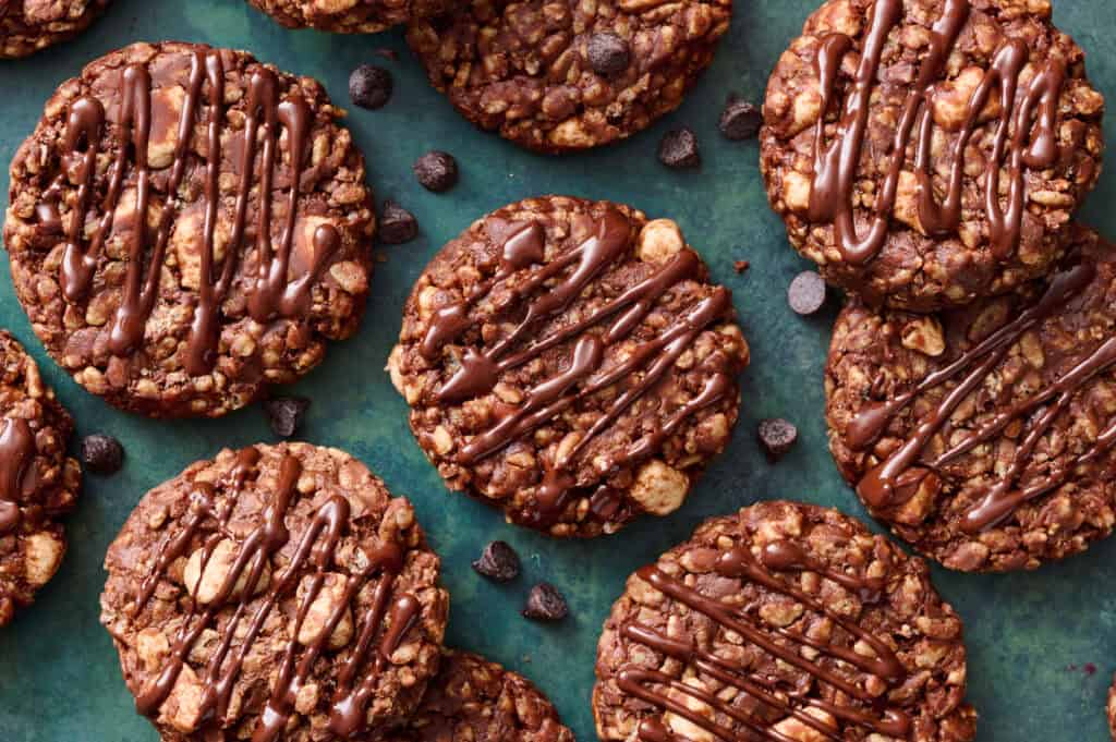 Chocolate oatmeal cookies with chunks of nuts, drizzled with chocolate, are arranged on a green surface with chocolate chips scattered around.