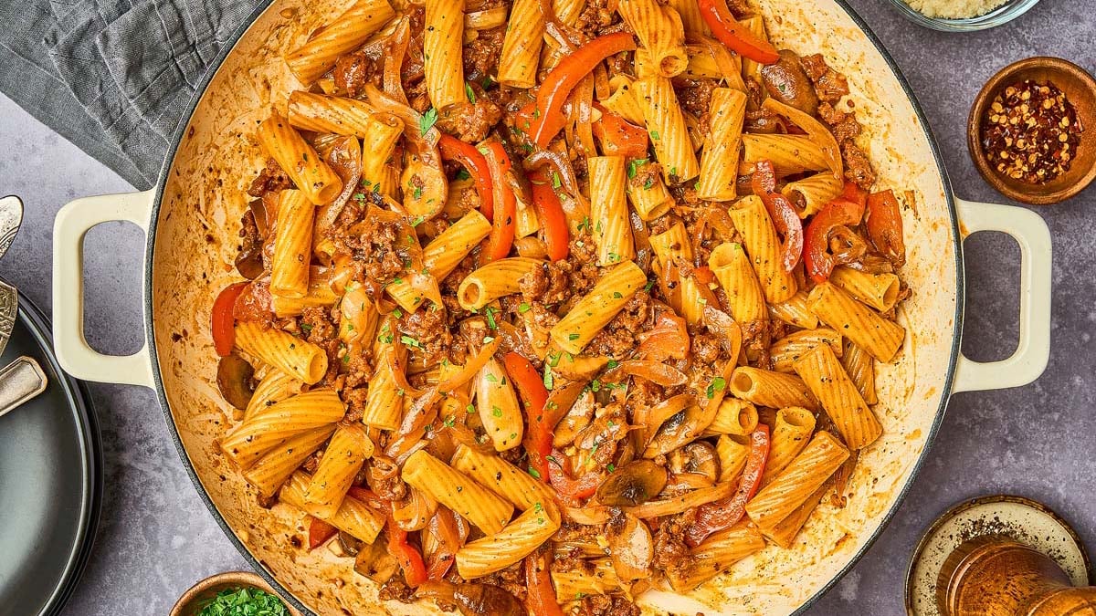 Large skillet filled with rigatoni pasta mixed with ground meat, bell peppers, mushrooms, and tomato-based sauce. Plates and spices are arranged around the pan.