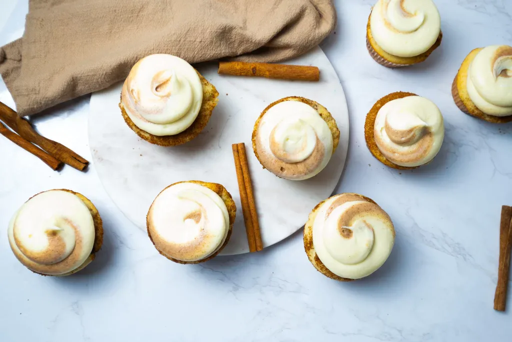 Cupcakes with swirled frosting and a sprinkle of cinnamon are arranged on a white surface with a round marble board, cinnamon sticks, and a beige cloth.
