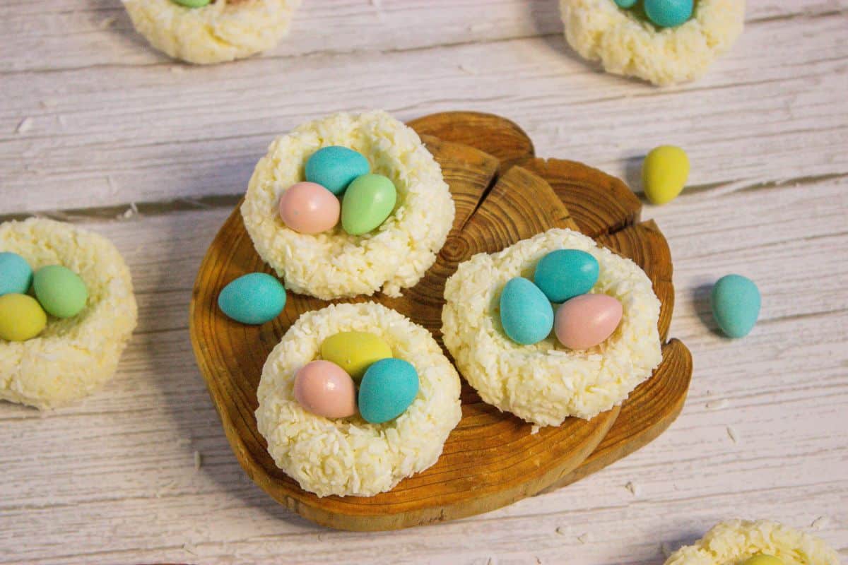 Round cookies topped with shredded coconut are shaped like nests and filled with pastel-colored candy eggs, displayed on a wooden board and white surface.