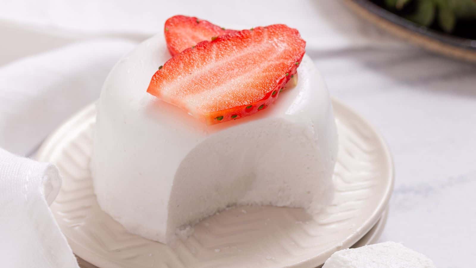 A dome-shaped white dessert with a slice removed, topped with two strawberry slices, is presented on a small plate.