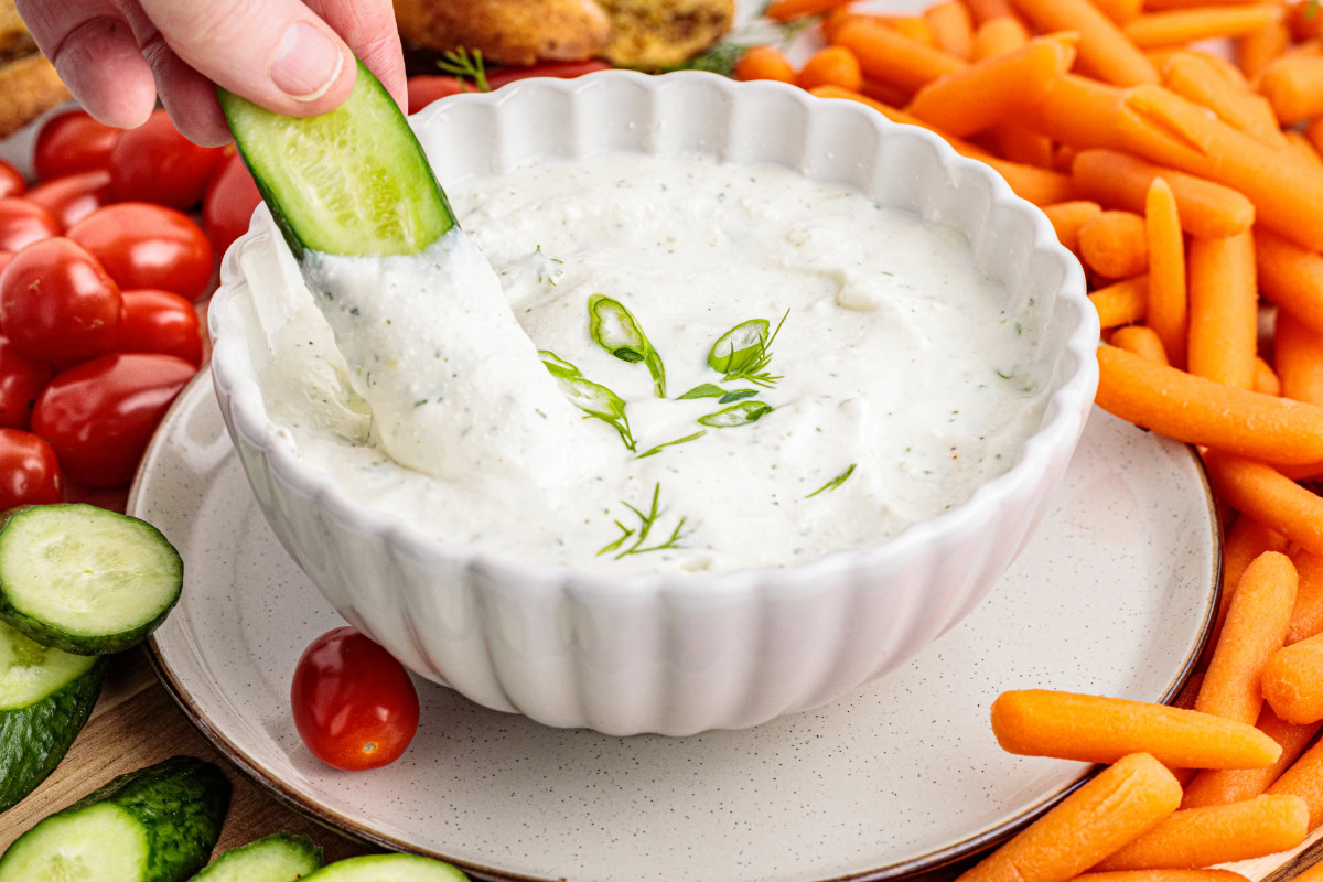 A hand dips a cucumber slice into a bowl of creamy white ranch dip, surrounded by baby carrots, cherry tomatoes, and more cucumber slices.