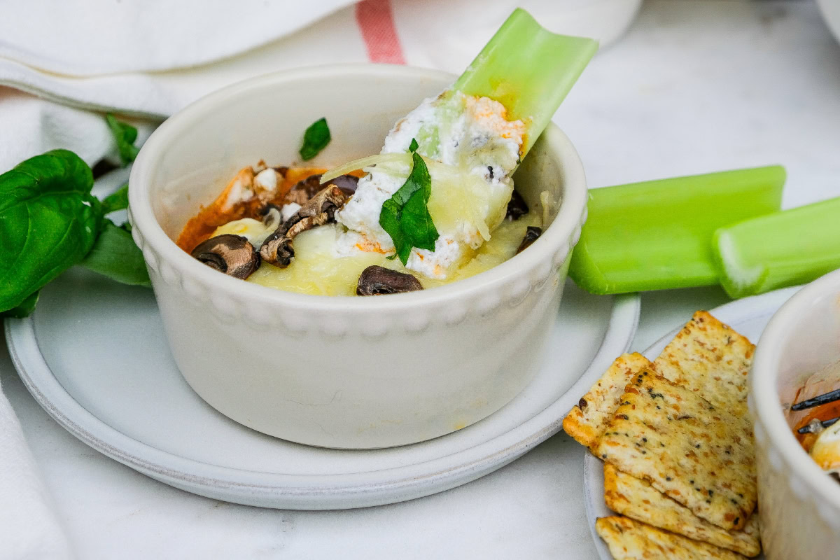 A white ramekin filled with a cheesy dip, garnished with mushrooms and herbs, with a celery stick dipped inside and crackers on the side.