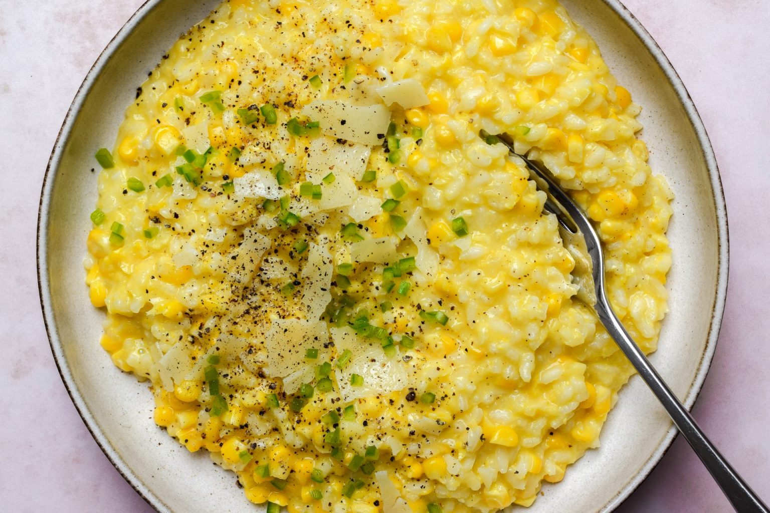 A plate of creamy corn risotto topped with chopped green herbs, grated cheese, and black pepper, with a fork resting on the side.