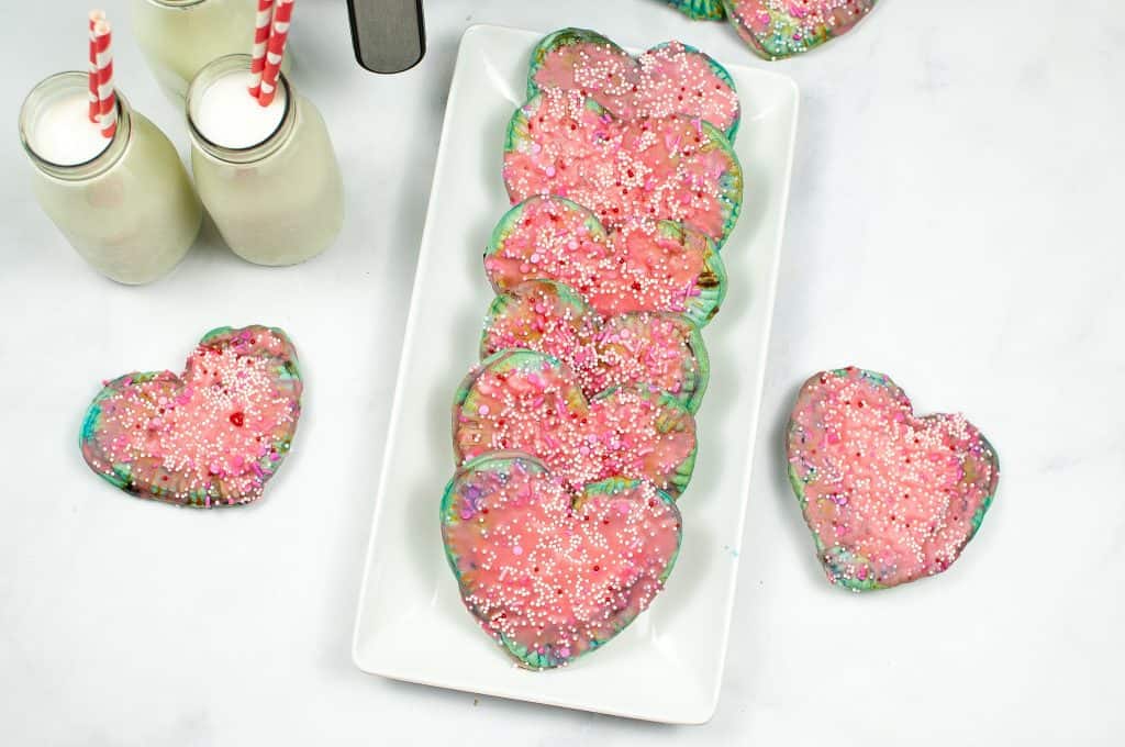 A rectangular white plate with heart-shaped cookies topped with pink icing and sprinkles, next to two bottles of milk with striped straws on a white surface.