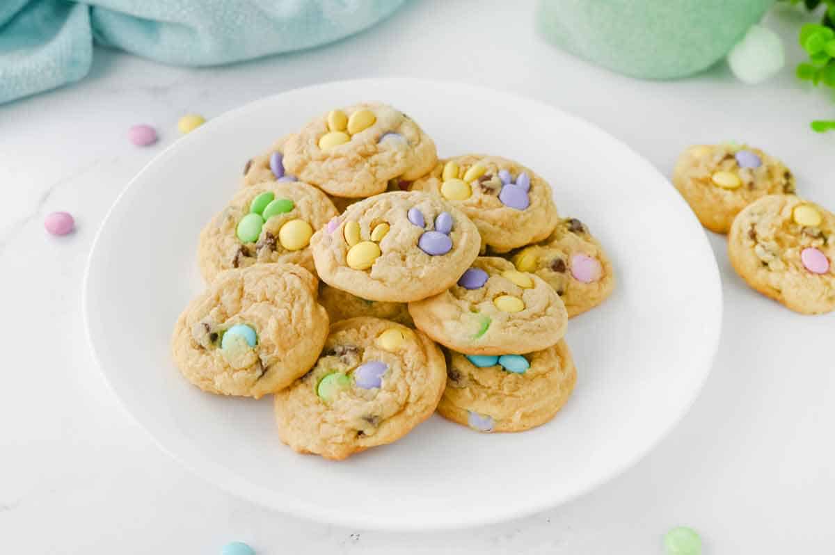 A white plate holds a pile of cookies topped with pastel-colored chocolate candies; a few cookies and candies are scattered on the white surface nearby.