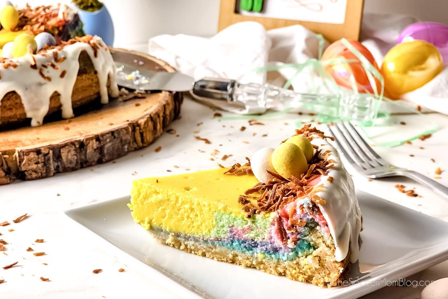 A slice of colorful layered cheesecake with white icing, chocolate shavings, and candy eggs on a plate; whole cake and utensils in the background.