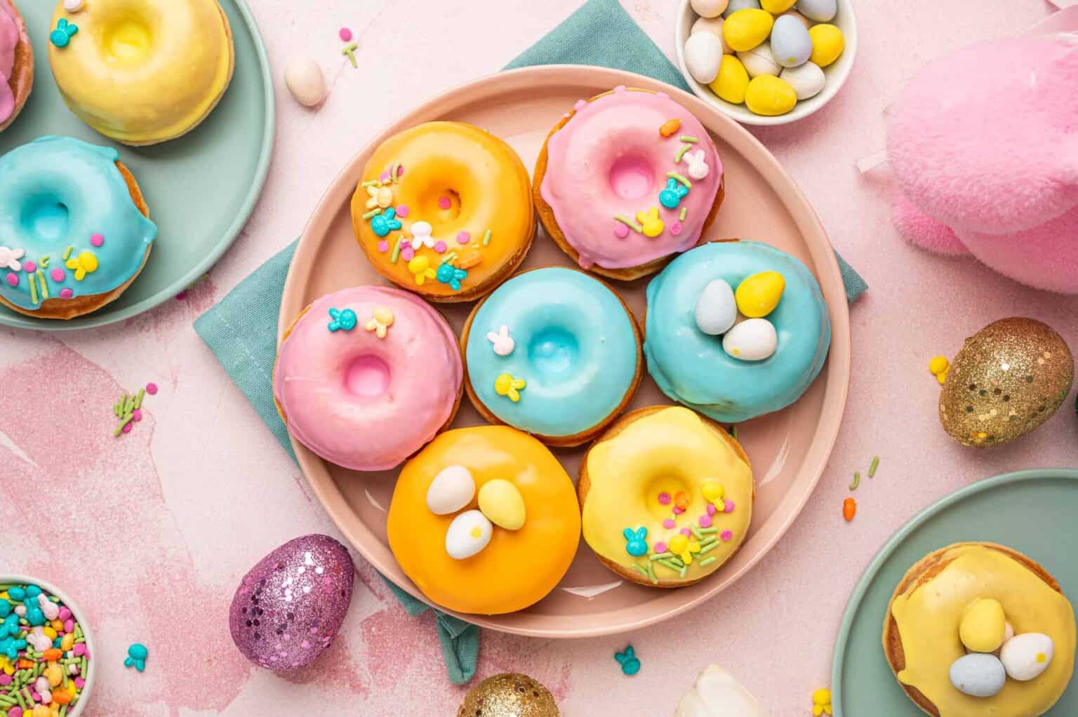 A plate of pastel frosted donuts topped with sprinkles and candy eggs, surrounded by bowls of candies and Easter eggs on a pink surface.
