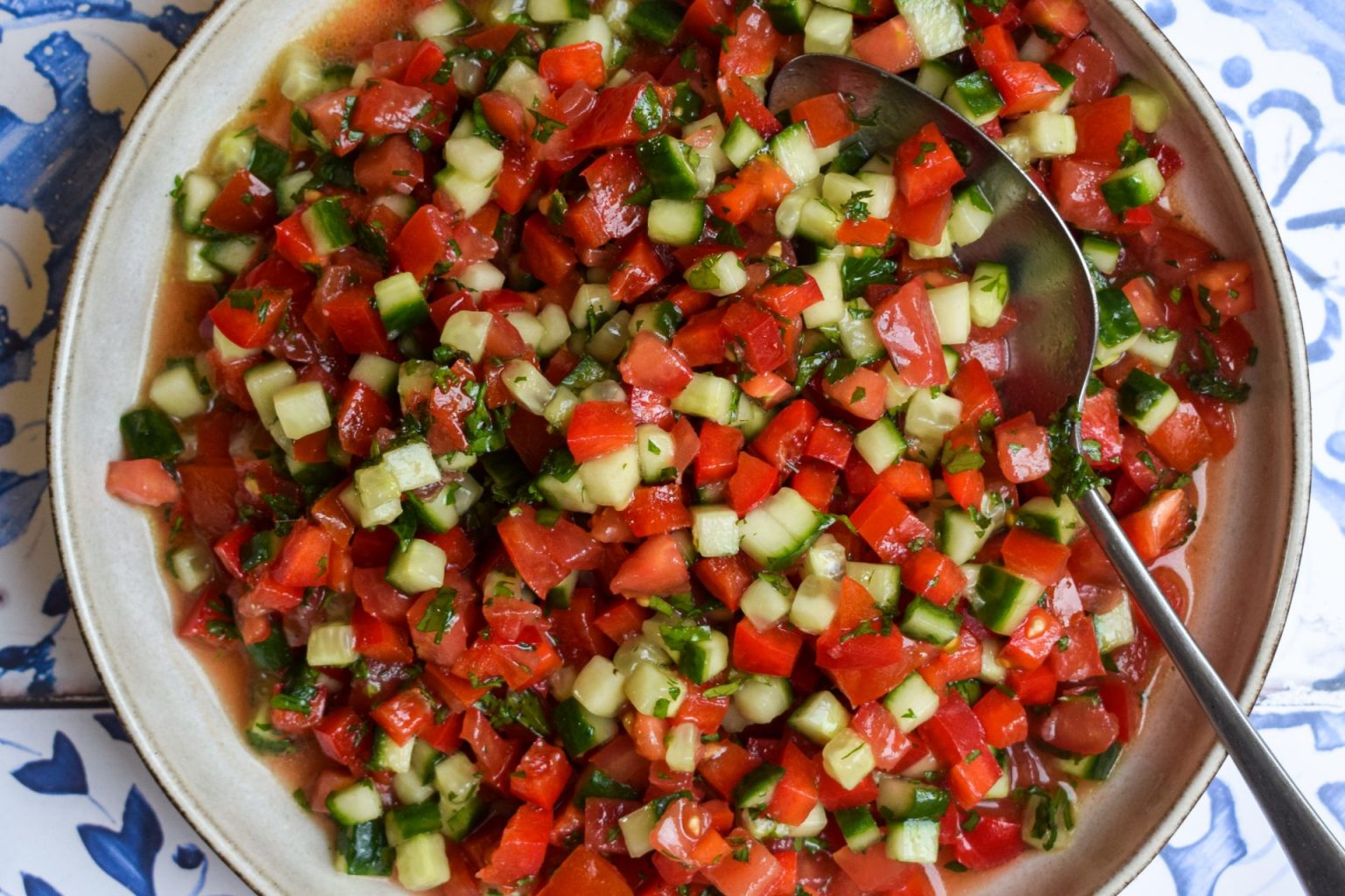 A bowl of chopped salad with diced tomatoes, cucumbers, and herbs, with a spoon resting inside the bowl, placed on a patterned surface.