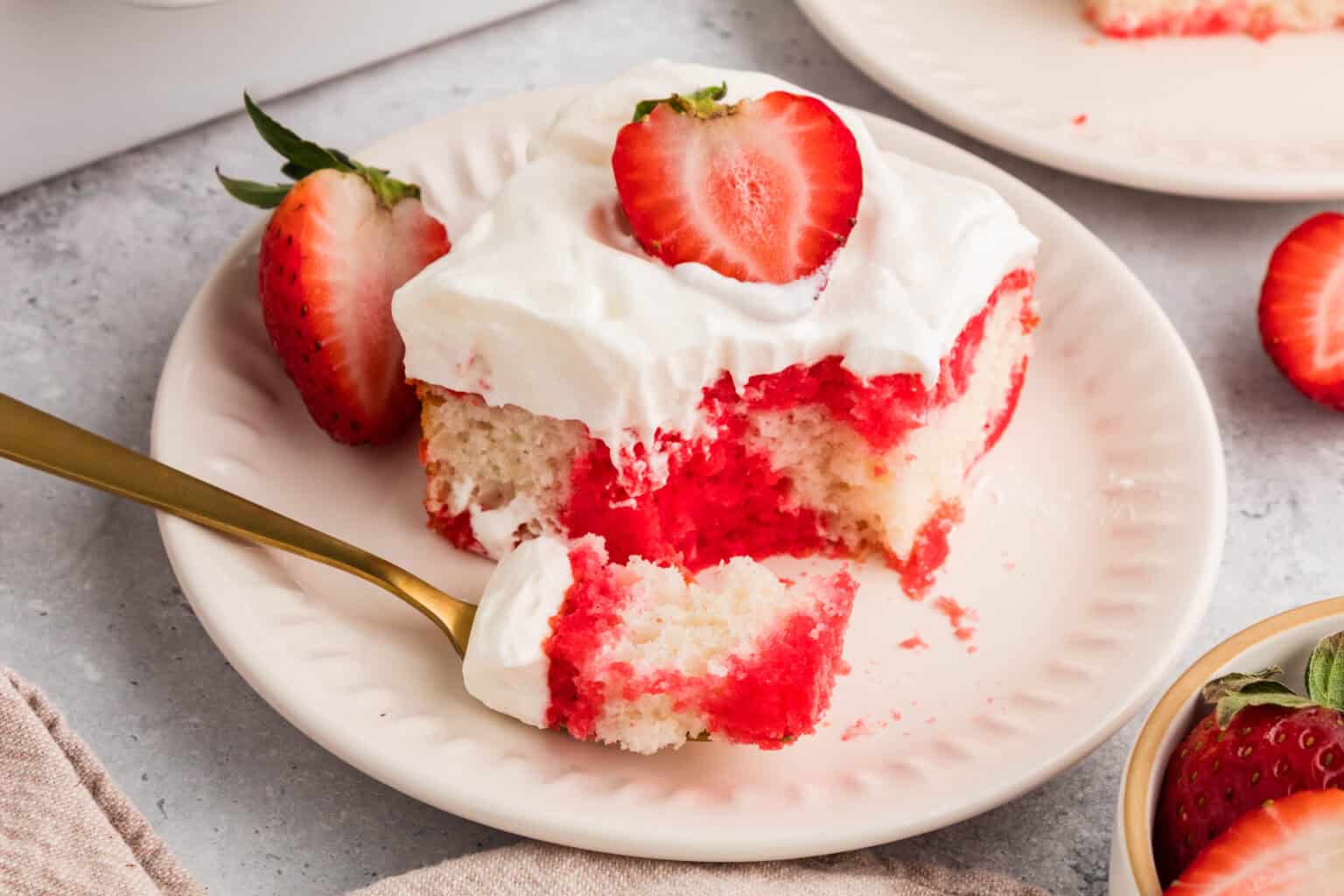 A slice of strawberry poke cake topped with whipped cream and a halved strawberry on a white plate, with a fork holding a bite-sized piece.