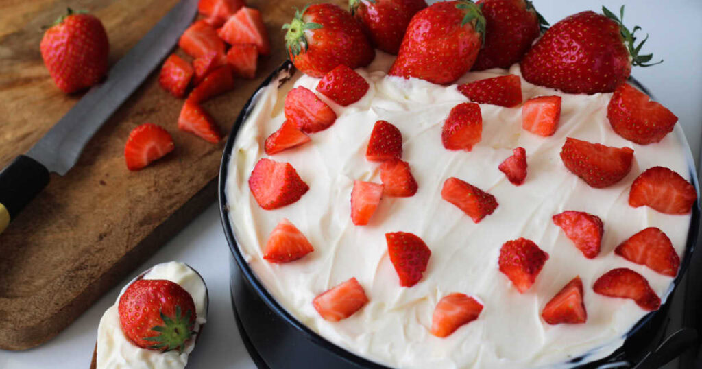 A cheesecake topped with whole and sliced strawberries sits next to a cutting board with a knife, strawberries, and a spoonful of cheesecake.