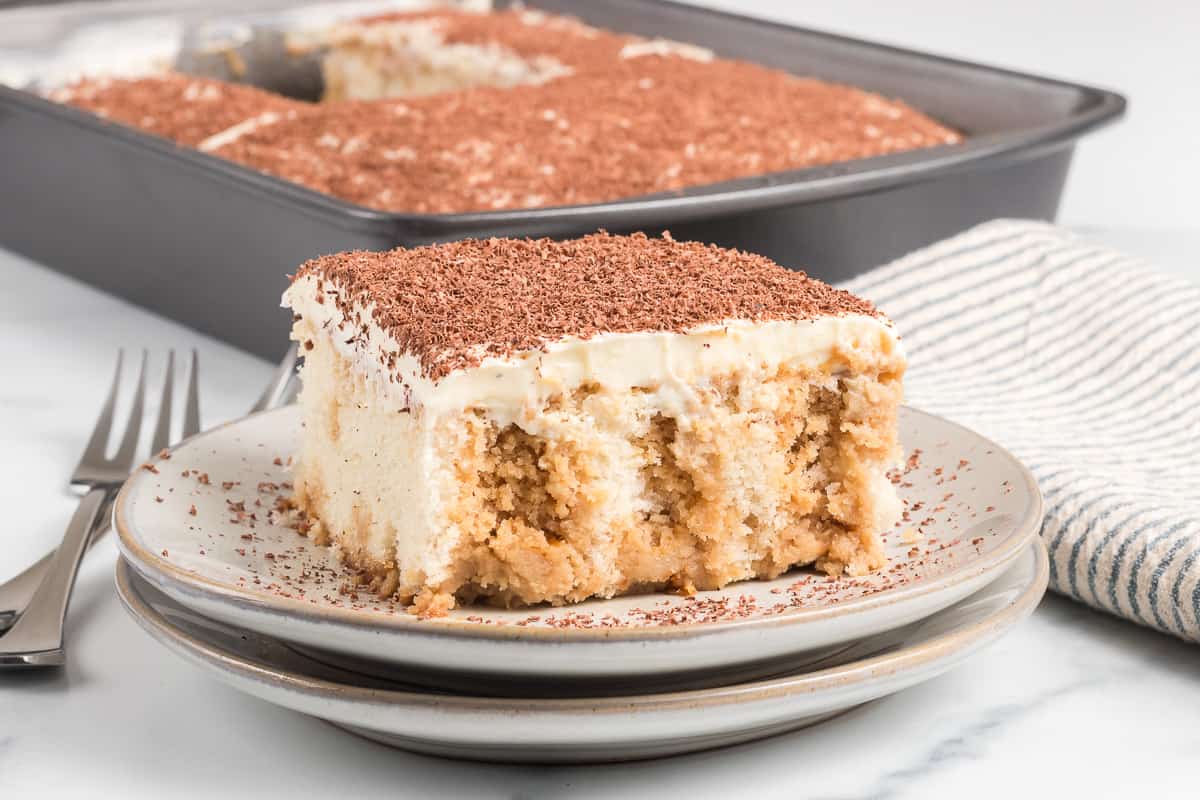 A slice of tiramisu poke cake topped with grated chocolate is served on a plate, with the remaining cake in a baking pan in the background.