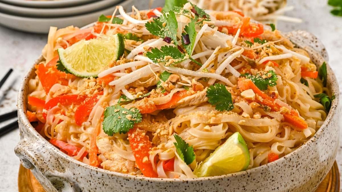 A bowl of Pad Thai noodles topped with bean sprouts, cilantro, crushed peanuts, lime wedges, and sliced vegetables.