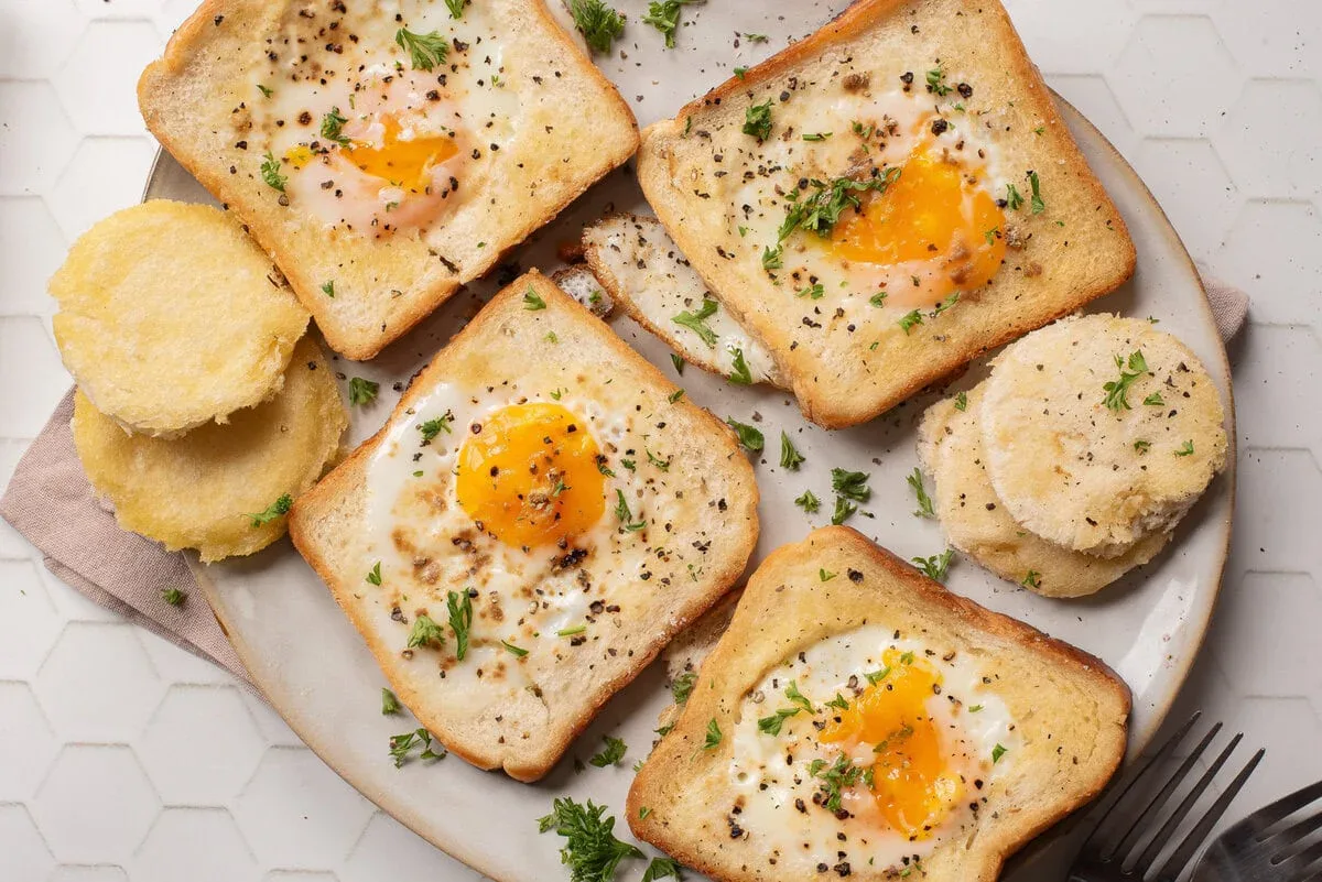 Four slices of toast with eggs cooked in the center are on a plate, garnished with black pepper and parsley, alongside round cutouts of toasted bread.