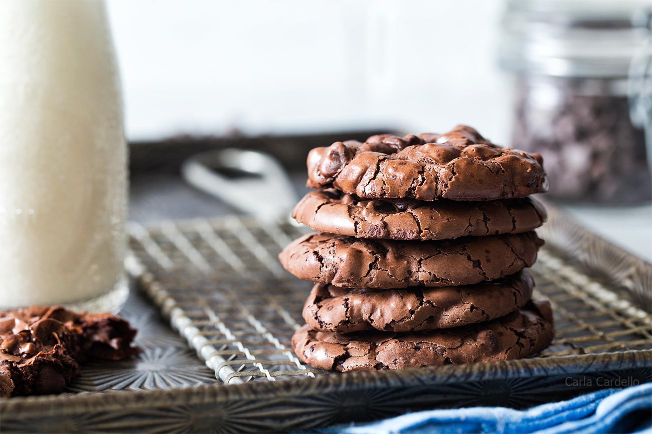 A stack of five chocolate cookies sits on a cooling rack next to a bottle of milk, with a jar of chocolate chips and a broken cookie in the background.