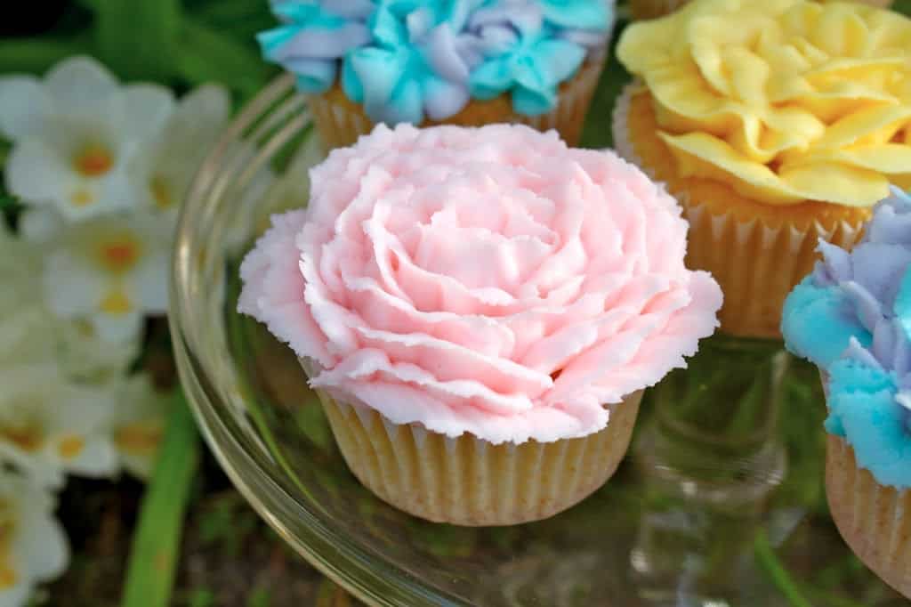 A cupcake topped with pink frosting shaped like a flower sits on a glass plate with other cupcakes decorated in blue and yellow flower designs.
