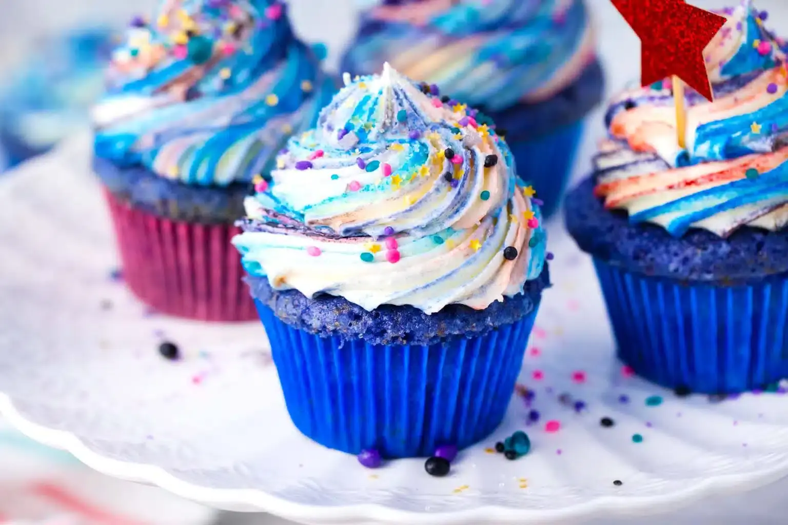 Colorful cupcakes with blue and purple swirled frosting, multicolored sprinkles, and blue wrappers arranged on a white plate.