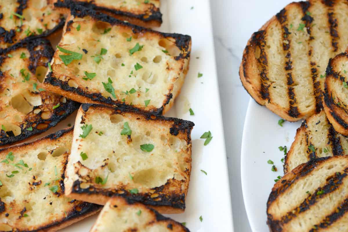 Slices of grilled bread, some charred, topped with chopped herbs, arranged on white plates.