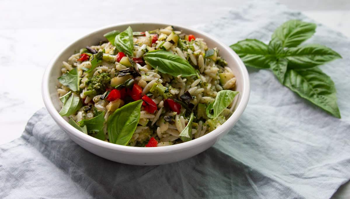 A white bowl filled with orzo pasta salad containing diced vegetables and fresh basil leaves, placed on a light blue cloth with basil leaves nearby.