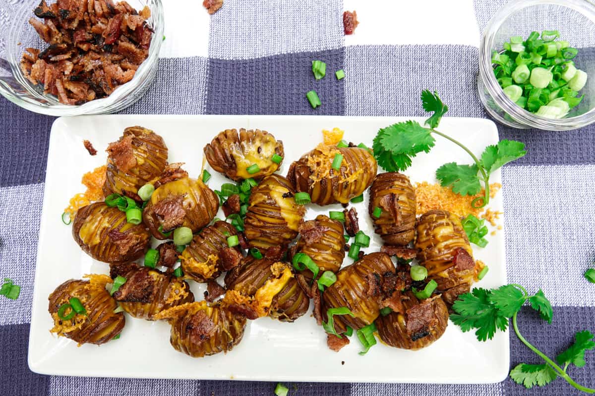 A rectangular white plate with sliced roasted potatoes topped with chopped green onions and bacon, garnished with cilantro, on a checkered cloth. Small bowls with toppings are nearby.