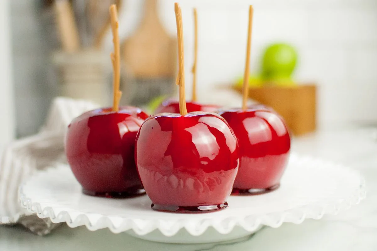 Four bright red candy apples with wooden sticks are displayed on a white scalloped-edged plate.