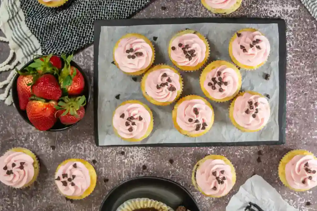 A tray of cupcakes with pink frosting and chocolate chips, surrounded by a bowl of strawberries and additional cupcakes on a textured surface.