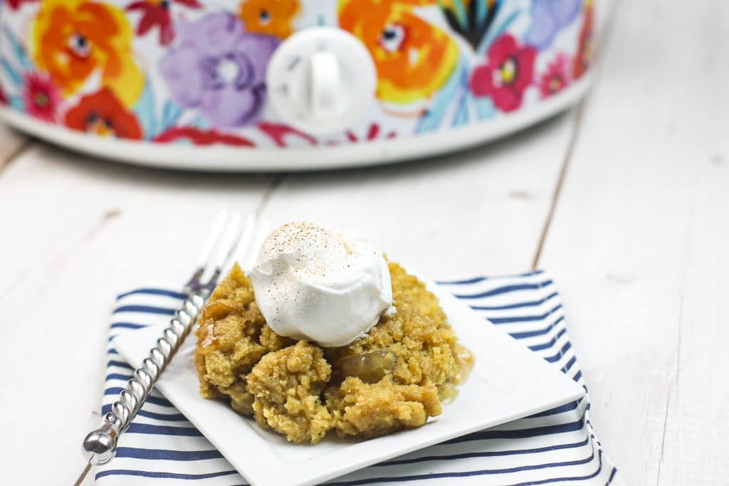 A plate of apple crumble topped with a scoop of whipped cream sits on a striped napkin, with a fork and a floral slow cooker in the background.