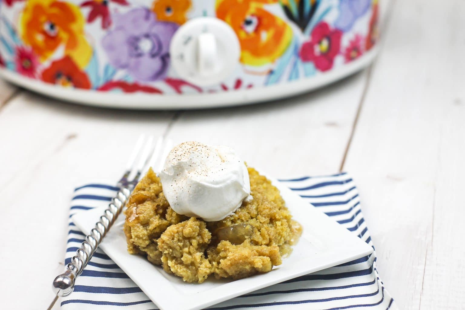 A serving of apple dessert topped with whipped cream sits on a white square plate with a striped napkin and fork; a floral slow cooker is in the background.