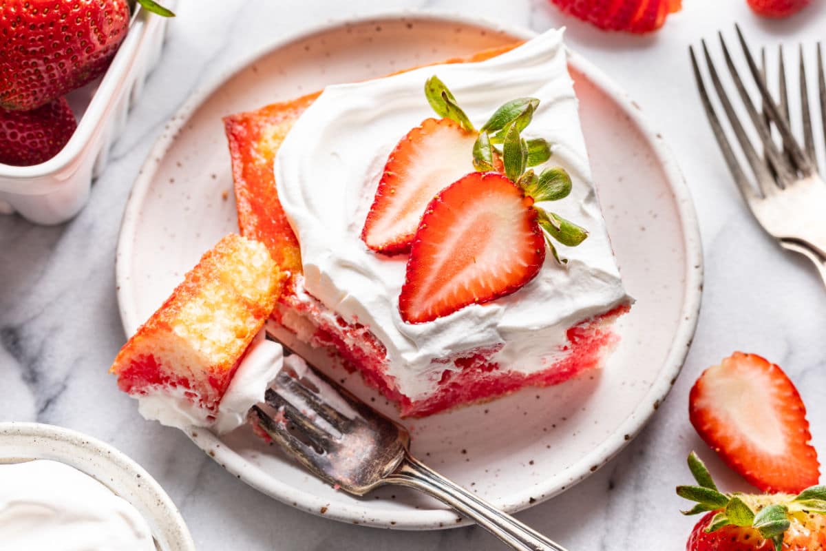 A slice of strawberry poke cake with whipped cream and fresh strawberry slices on a plate, with a fork holding a bite.