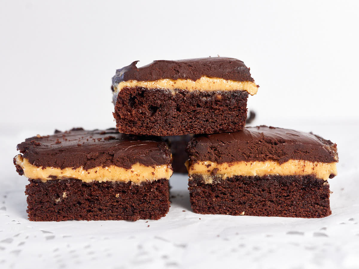 Three stacked chocolate brownies with a peanut butter layer and chocolate frosting, displayed on a white surface.