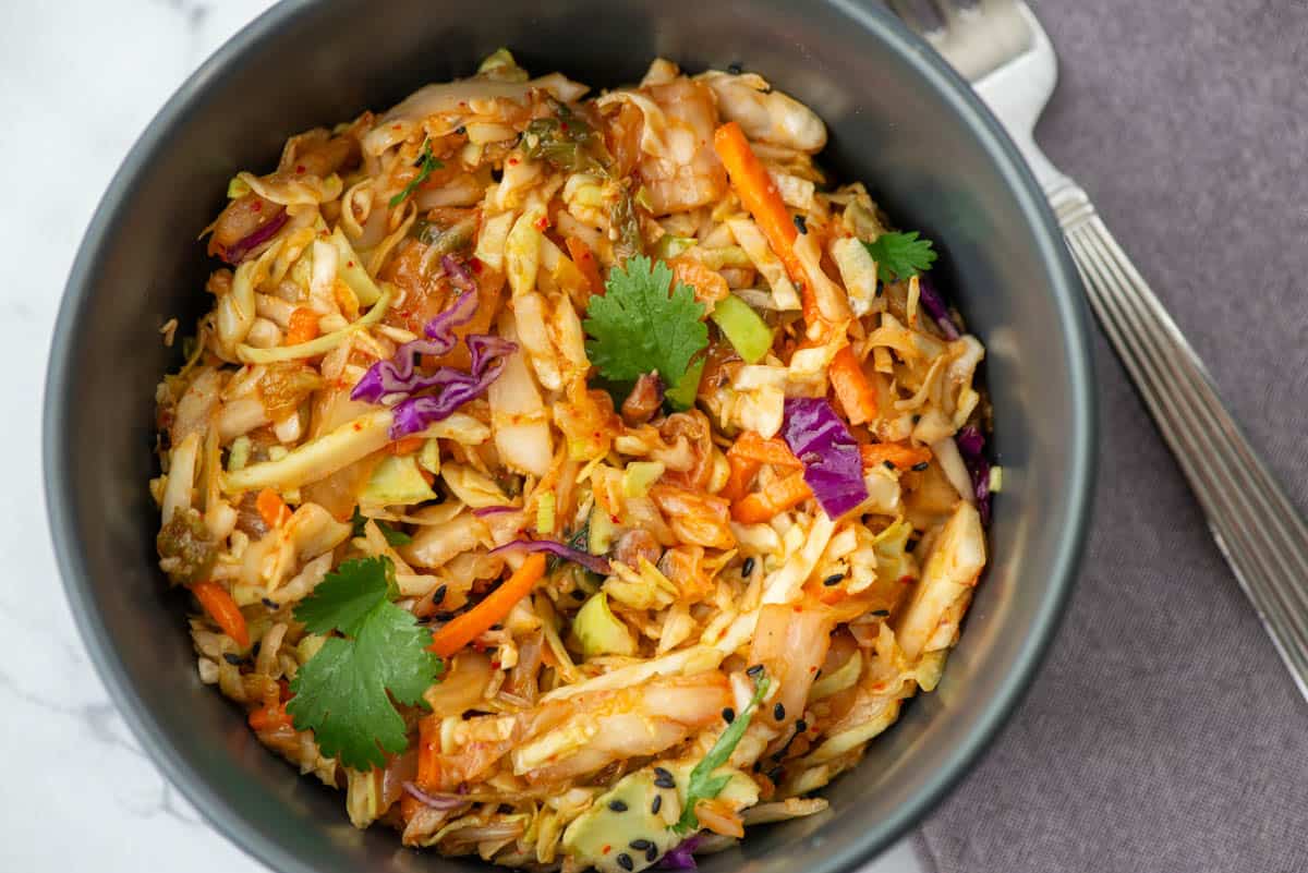 A bowl of stir-fried cabbage, carrots, and red cabbage garnished with cilantro, served with a fork on the side.