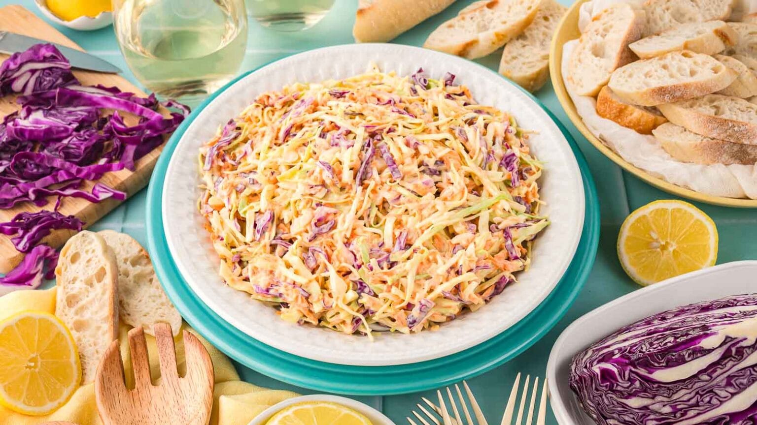 A bowl of coleslaw made with shredded green and purple cabbage, surrounded by sliced bread, lemon halves, and cabbage wedges on a table.