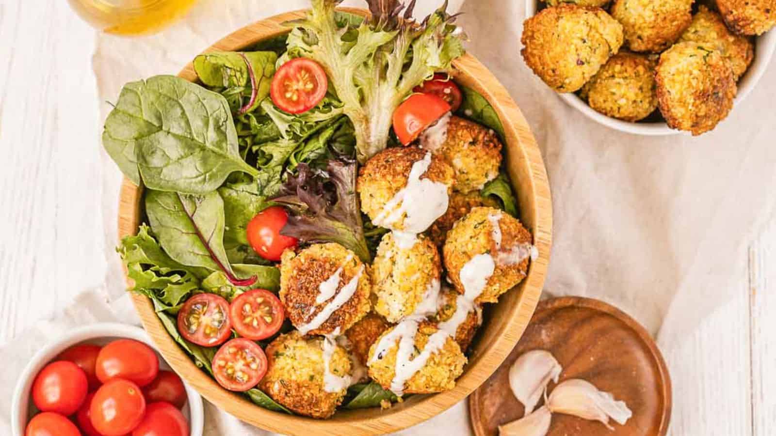 A wooden bowl filled with mixed greens, cherry tomatoes, and falafel drizzled with sauce. A bowl of cherry tomatoes and a bowl of falafel are also nearby.