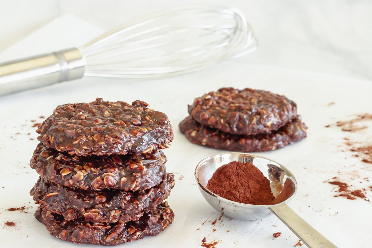 A stack of chocolate oatmeal cookies next to a small pile of cookies, a metal whisk, and a measuring spoon filled with cocoa powder on a white surface.