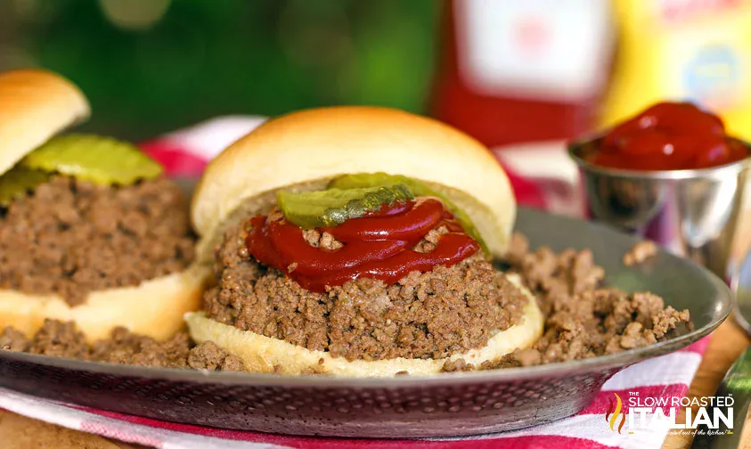 A sloppy joe sandwich topped with ketchup and pickle slices served on a bun, with ground beef spilling onto the plate.