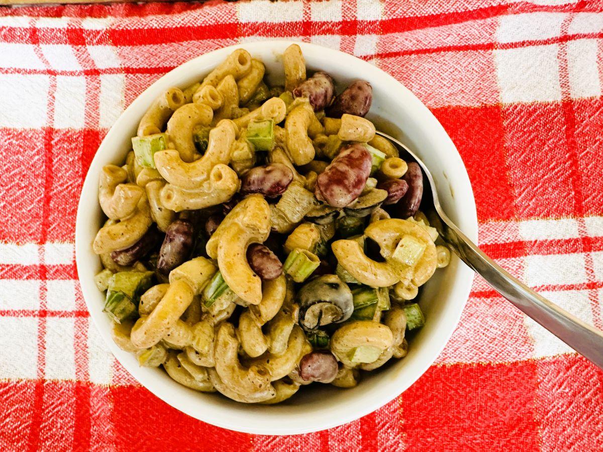 A white bowl of macaroni salad with kidney beans, celery, and a creamy dressing, placed on a red and white checkered cloth. A metal spoon rests inside the bowl.