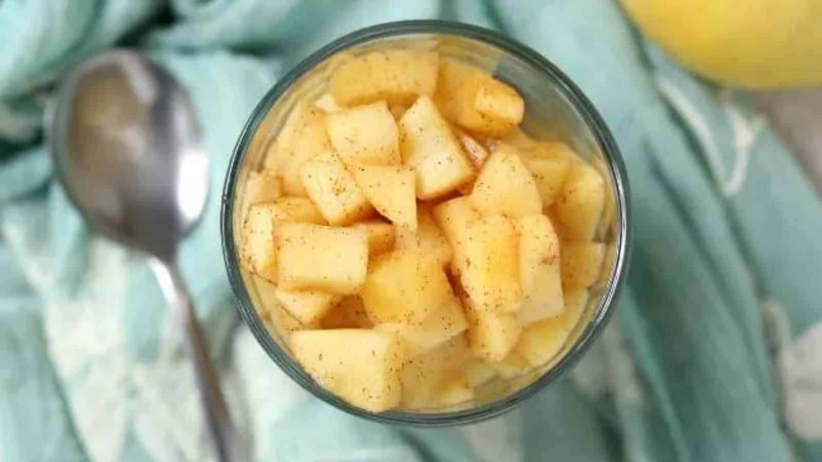 A glass filled with diced apples sprinkled with cinnamon, placed on a light blue cloth next to a spoon.
