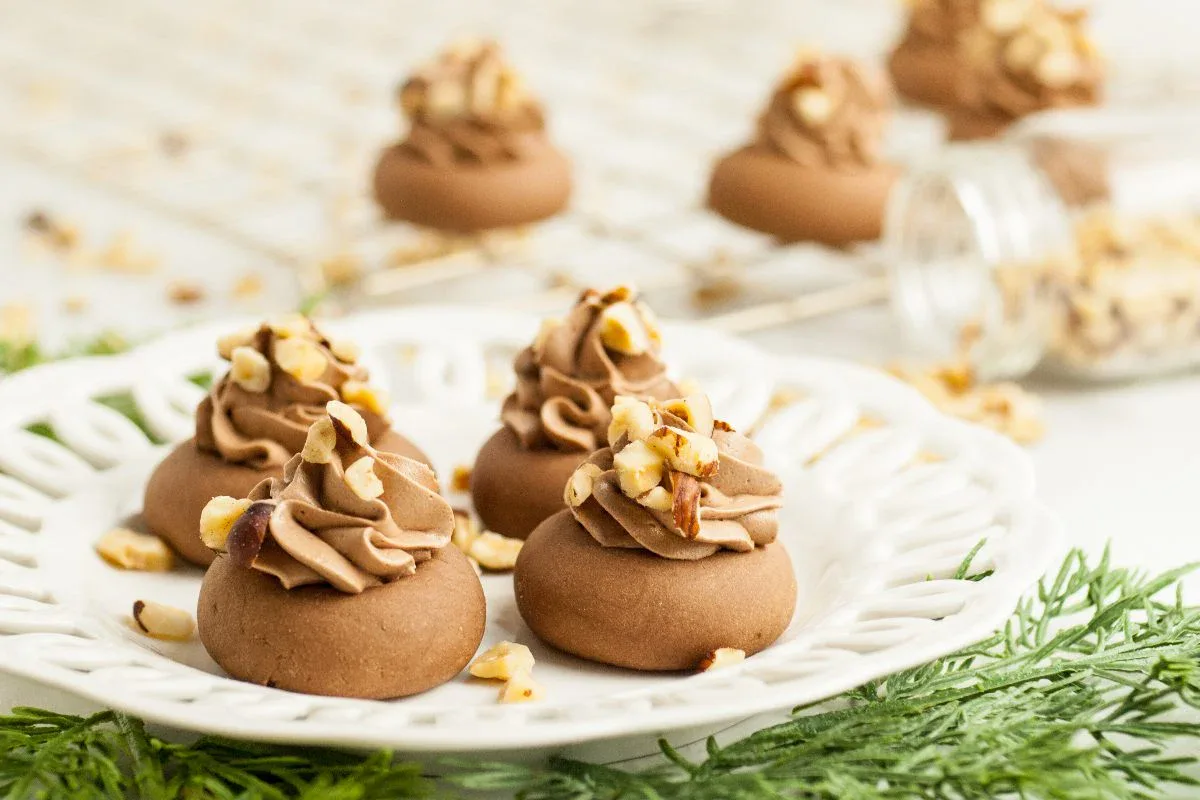 A white plate with chocolate cookies topped with chocolate frosting and chopped nuts, with more cookies and a jar of nuts in the background.