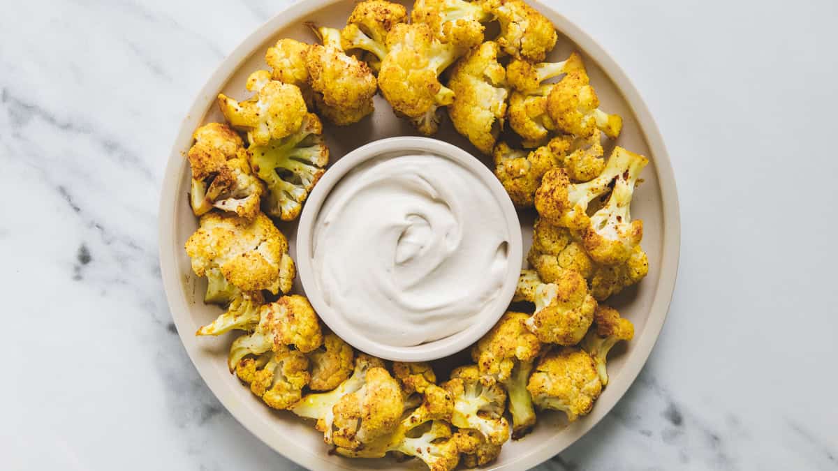 A plate of roasted cauliflower florets arranged around a bowl of creamy white dipping sauce on a marble surface.