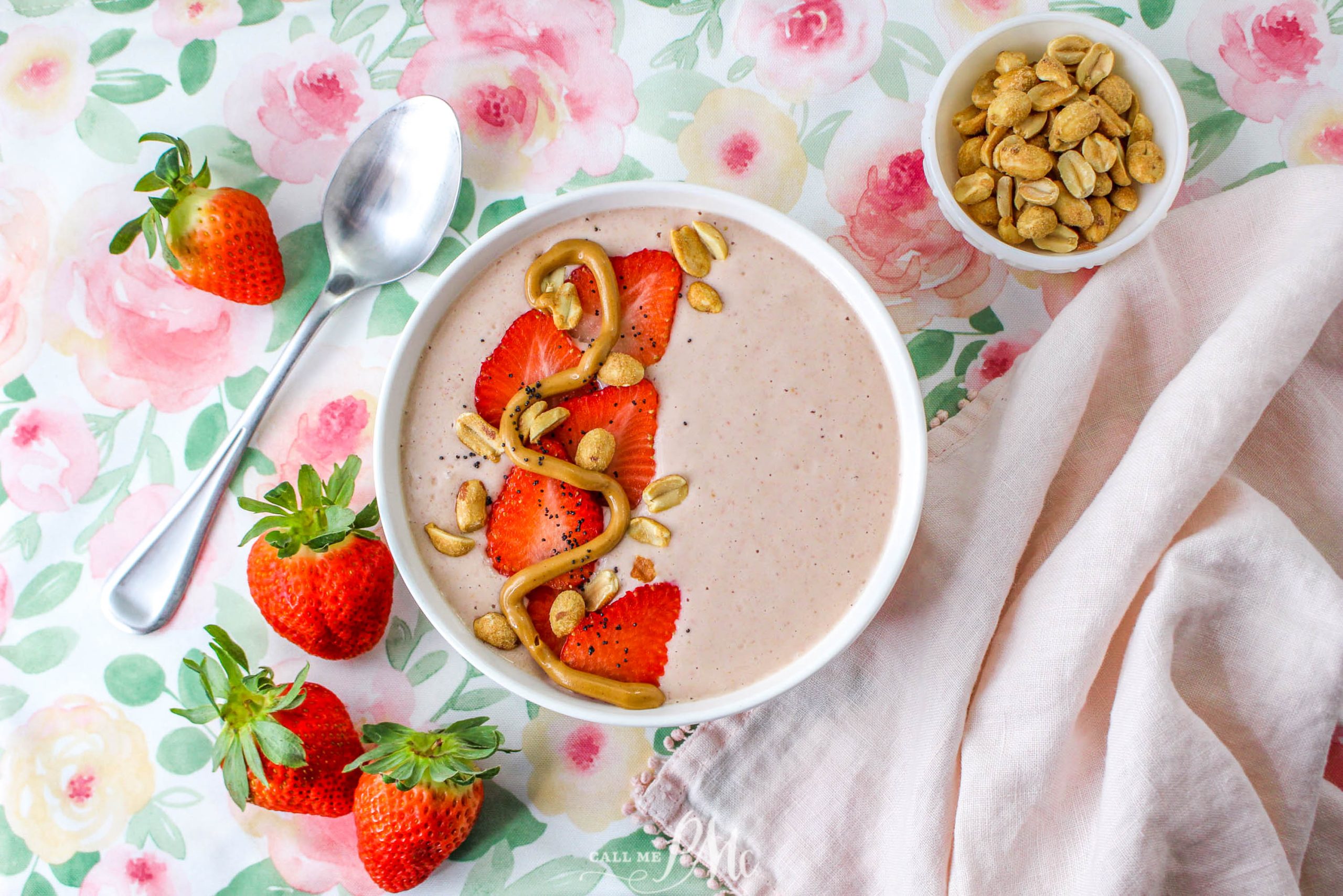 A smoothie bowl topped with sliced strawberries, peanuts, and a drizzle of peanut butter, next to a spoon, whole strawberries, and a small bowl of peanuts on a floral tablecloth.